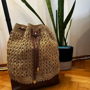 May include: A handcrafted, tan crochet drawstring bag with a dark brown leather base and straps. The bag features a unique woven pattern and gold-colored accents. The bag is a stylish accessory.