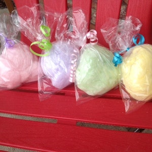 May include: Four bags of cotton candy in different colors: pink, purple, green, and yellow. The bags are tied with colorful ribbons and are sitting on a red bench.