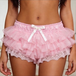 Puede incluir: Pantalones cortos rosas con volantes y lazo blanco. Los pantalones cortos presentan múltiples capas de tejido transparente, creando un efecto en capas. El borde inferior está adornado con encaje blanco. Los pantalones cortos son de un material suave y sedoso.