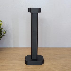 Puede incluir: Un soporte negro para auriculares con una base rectangular y un soporte vertical alto. La parte superior del soporte tiene una sección más ancha para sujetar los auriculares. Una pequeña planta en maceta está a la izquierda del soporte sobre una superficie de madera.
