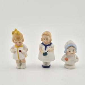May include: Three small porcelain figurines of children. The left figure wears a white dress with yellow accents and a yellow bow. The center figure wears a white dress with a blue sailor collar and a green bag. The right figure wears a blue bonnet.