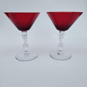 May include: Two martini glasses featuring deep red, conical bowls and clear, textured stems with a pineapple-like pattern. Each glass has a wide, circular clear base, providing stability for the vibrant red glass top section.