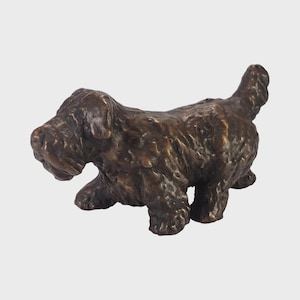 May include: A bronze dog sculpture, depicted in a walking stance. The dog features a textured coat and a short tail. The sculpture is a dark brown hue, set against a white backdrop.