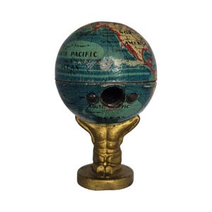 Op de afbeelding: Vintage globevormige puntenslijper. De globe toont een kaart met continenten en oceanen in blauw- en groentinten. Hij staat op een goudkleurige, sierlijke voet. De puntenslijper heeft twee gaten voor potloden.