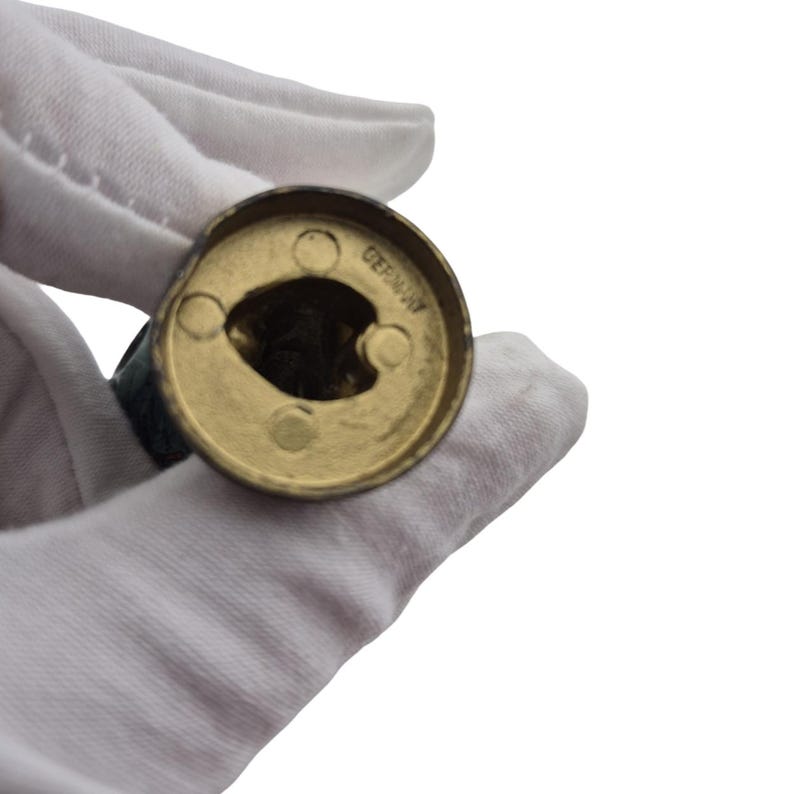 Op de afbeelding: Een close-up van een messingkleurig object met het woord "GERMANY" erop gestempeld. Het object heeft een cirkelvorm met een centrale opening en drie kleinere gaten. Het object wordt vastgehouden door een gehandschoende hand.