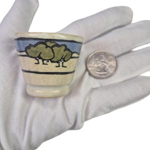 Puede incluir: Una pequeña taza de cerámica moteada con borde acampanado, que presenta un diseño pintado a mano de dos árboles verdes sobre una banda azul claro, acentuado por finas líneas horizontales negras. Una moneda de 25 centavos está al lado.