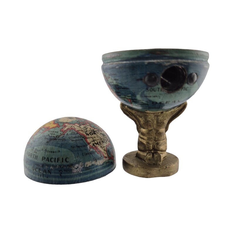 Op de afbeelding: Een vintage globe-vormige puntenslijper met een blauw en goud afwerking. De globe is in twee&euml;n gesplitst, waardoor het interne mechanisme zichtbaar wordt. De basis is een goudkleurige figuur. De globe toont de Noordelijke Stille Oceaan en Noord-Amerika.
