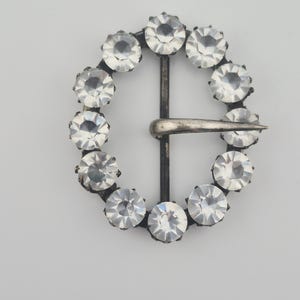 May include: A vintage silver-tone buckle with a circular design. The buckle is adorned with clear, faceted gemstones set around the perimeter. A central bar and pin complete the design, creating a classic and elegant accessory.