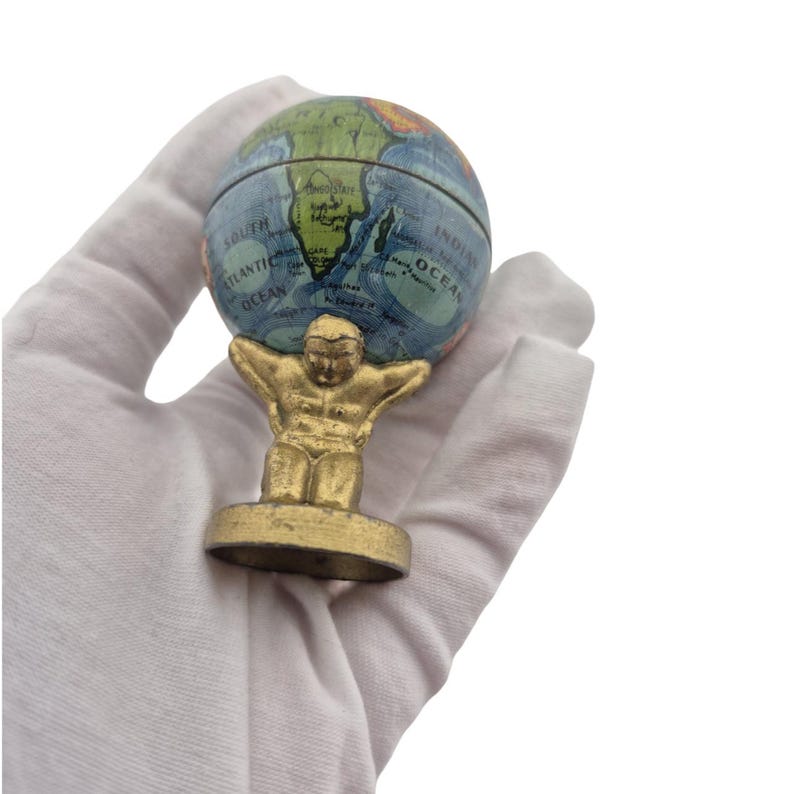 Op de afbeelding: Een miniatuur wereldbol, vastgehouden door een goudkleurige Atlas figuur. De wereldbol toont een kaart met blauwe oceanen en continenten in groen, geel en bruin. De Atlas figuur staat op een cirkelvormige basis.