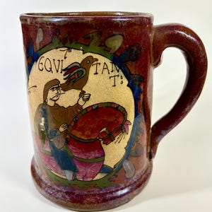 May include: A ceramic mug with a dark red glaze and a large handle. The mug features a central illustration within an oval frame, depicting a figure with a bird and a horse. The text "EQUI TANT" is visible. The mug's design has a medieval aesthetic.