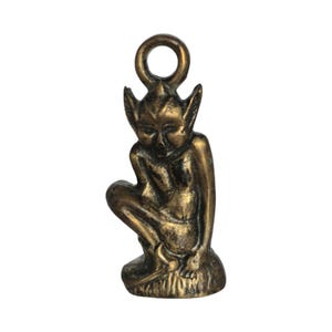 Könnte beinhalten: Ein bronzefarbener Metallanhänger, der eine sitzende Fee oder einen sitzenden Elf mit spitzen Ohren darstellt. Der Anhänger hat oben eine Schlaufe zum Befestigen an einer Halskette oder einem Armband. Die Fee sitzt auf einem kleinen Sockel.