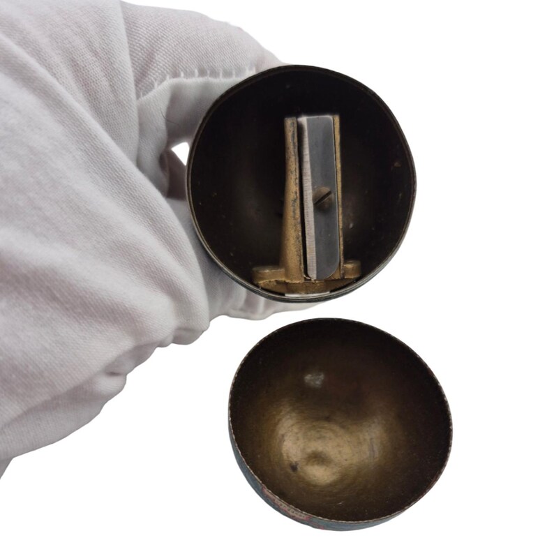 Op de afbeelding: Een vintage puntenslijper met een messingkleurige metalen behuizing en een zilverkleurig mes. De puntenslijper is rond met een afneembaar deksel, dat het mes en de binnenkant onthult. Deksel en behuizing vertonen tekenen van ouderdom en gebruik.