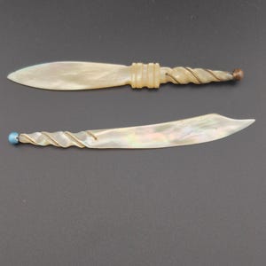 以下が含まれることがあります： 虹色に輝くマザーオブパール製のレターオープナー2本。上のオープナーは直線の刃、リブ付きの持ち手、茶色の端が付いたねじれたグリップが特徴です。下のオープナーは湾曲した刃、ねじれた持ち手、青いビーズの端が付いています。