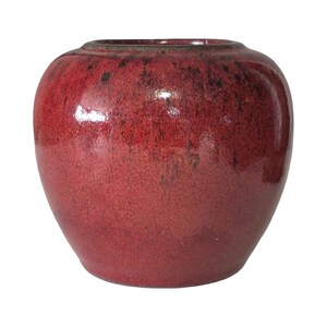 May include: A round, red ceramic vase with a glossy finish. The vase has a wide body and a slightly narrower opening at the top. The red color has darker, speckled accents.