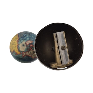 Op de afbeelding: Een vintage potloodslijper met een wereldbolvormige gum. De slijper heeft een donkere metalen behuizing, een zilveren mes en een messingkleurige basis. De wereldbolgum heeft een kaartontwerp met blauwe, gele en rode tinten.