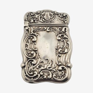 May include: A silver-toned metal vesta case featuring an ornate, embossed scrollwork design with floral motifs and a central shell-shaped element at the top. The rectangular container has a hinged lid and a smooth, polished finish on the front surface.