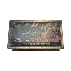 May include: A rectangular metal tin with a black and teal finish featuring gold-toned decorative illustrations of a person and floral motifs. The text reads Assortment Parisien L'incomparable and PARK & TILFORD NEW YORK PARIS on the front surface.