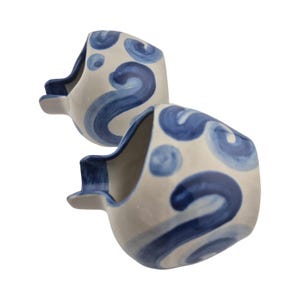 Puede incluir: Dos recipientes de cerámica con forma de pez. Los recipientes son blancos con diseños de olas y remolinos azules. Los recipientes tienen un asa pequeña y rectangular en un lado y una abertura curva en el otro.