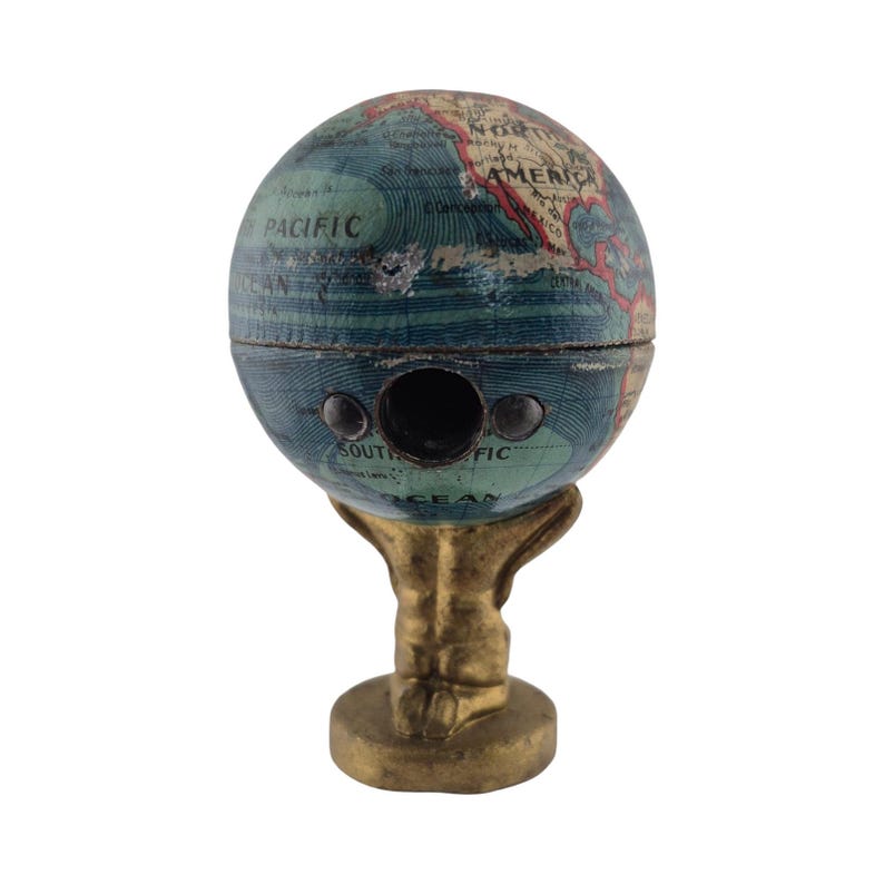 Op de afbeelding: Een vintage globe-vormige puntenslijper met een blauw en groen kaartontwerp. De puntenslijper heeft drie gaten en staat op een goudkleurige standaard. Het woord "PACIFIC" is zichtbaar op de globe.