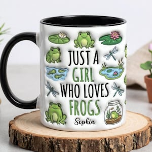 Op de afbeelding: Witte keramische mok met een zwarte handgreep en binnenkant, met de tekst "JUST A GIRL WHO LOVES FROGS" en een naam. De mok is versierd met groene kikkers, waterlelies, libellen en kleine vijvers. De mok staat op een houten onderzetter.