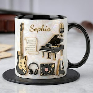 May include: White ceramic mug with black handle and rim, featuring musical instrument designs. Includes a guitar, piano, microphone, headphones, and a record player. The name "Sophia" is written in gold script on the mug.