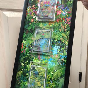 May include: A framed artwork with three Pokémon trading cards set against a jungle backdrop. The cards are in clear holders, and the frame is black. The artwork features a detailed illustration of a lush, green environment with colorful flowers and a small pond.