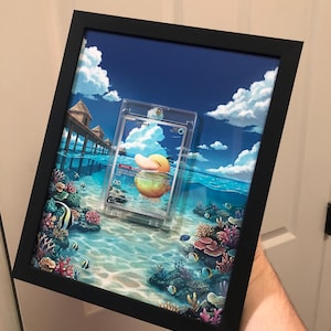 Puede incluir: Un marco rectangular negro tipo caja con una tarjeta coleccionable de Psyduck dentro de una funda protectora de plástico transparente. El fondo muestra una ilustración vibrante de un océano tropical con arrecifes de coral y peces.