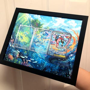 May include: A black rectangular frame containing a layered 3D shadow box art piece. The artwork features three Pokémon trading cards, Froakie, Frogadier, and M Greninja-EX, set against a vibrant, multi-layered underwater scene with blue water.