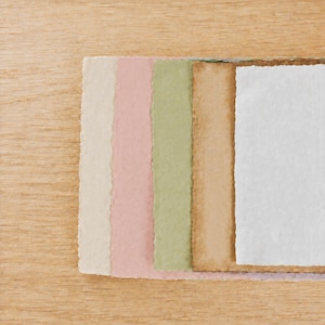 May include: A stack of five rectangular sheets of handmade paper with deckled edges, arranged in a cascading row. The colors include cream, soft pink, sage green, tan, and white, each featuring a distinct fibrous texture and uneven borders.