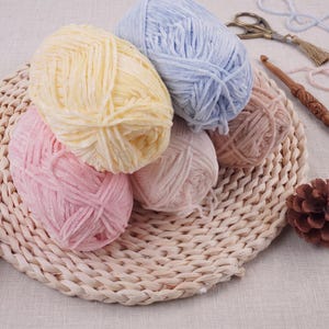 May include: Five skeins of soft, velvet-textured yarn in pastel shades of yellow, light blue, pink, and dusty rose. The yarn is arranged on a woven circular mat alongside a wooden crochet hook, a pair of metal scissors, and a small pinecone.