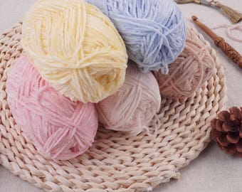 May include: Five skeins of soft, velvet-textured yarn in pastel shades of yellow, light blue, pink, and dusty rose. The yarn is arranged on a woven circular mat alongside a wooden crochet hook, a pair of metal scissors, and a small pinecone.
