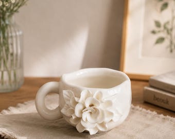 May include: A white ceramic teacup with a sculpted floral design on the side. The cup has a rounded handle and sits on a beige linen runner. A vase of white flowers and framed botanical art are in the background.