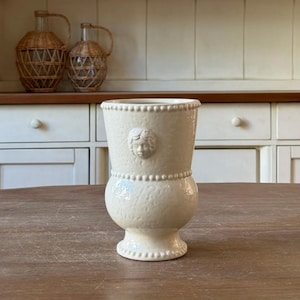 May include: Off-white ceramic vase with a textured surface and a classical face relief. The vase has a bulbous base, a cylindrical body, and a beaded rim. It is displayed on a wooden table.