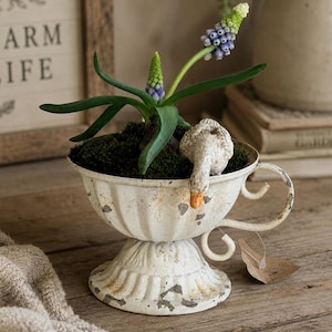 May include: A white metal cup-shaped planter with a distressed finish, featuring a small decorative goose figurine perched on the rim. The planter contains dark green moss and a flowering grape hyacinth plant with blue and green buds.