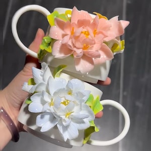 May include: Two white ceramic mugs with large, 3D floral embellishments. One mug features a pink flower, the other a white flower, both with yellow centers and green leaves. The mugs have curved handles.