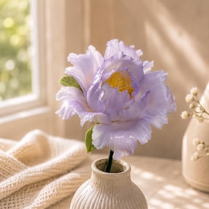 May include: A single artificial peony flower with ruffled, light purple petals and a bright yellow centre. The flower features small green leaves attached to a thin stem, which is placed inside a small, ribbed, off-white ceramic vase.