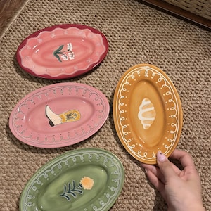 以下が含まれることがあります： ピンク、オレンジ、緑の色合いの、4つの楕円形の装飾用セラミックプレート。各プレートには、花柄やブーツのモチーフなど、ユニークな手描きのデザインが施されています。プレートは織り込まれた表面に配置されています。