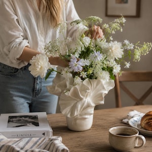 Könnte beinhalten: Eine weiße Keramikvase mit einem gerüschten, blütenblattartigen Rand, gefüllt mit einem Strauß aus weißen Pfingstrosen, grüner Wilde Möhre und hellvioletten Blumen. Die Vase hat eine glatte, glänzende Oberfläche und eine weit ausgestellte Öffnung.