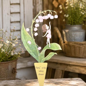 Puede incluir: Un estaca decorativa de jardín con una ilustración estilo acuarela de un hada de pie sobre una hoja verde bajo un racimo de flores de lirio de los valles rosa pálido. La base triangular amarilla muestra el texto Fairy's lily valley dream.