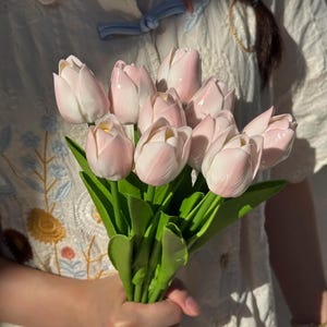 Może przedstawiać: Bukiet jasno-różowych i białych tulipanów z zielonymi liśćmi. Tulipy trzymane są w dłoni. Tło to biała koszula.