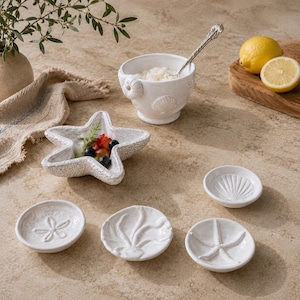 Könnte beinhalten: Eine Sammlung weißer Keramik-Servierschalen mit Küstenmotiv. Enthält eine sternförmige Schale, kleine runde Schalen mit Muscheldesign und eine Schüssel mit einem Löffel. Eine Zitrone und eine Schüssel Reis sind ebenfalls vorhanden.