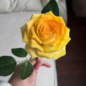 May include: A vibrant yellow rose with a gradient of color from light yellow to a deeper orange in the center. The rose has multiple petals and is accompanied by green leaves and a stem.