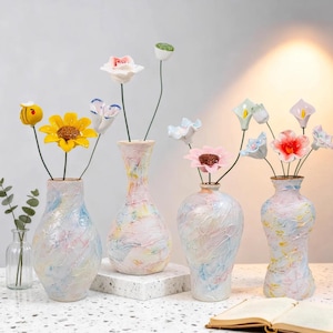 May include: A selection of decorative vases with marbled exteriors in pastel shades, each displaying artificial flowers. The flowers include sunflowers, calla lilies, and other blooms. The vases are arranged on a white surface.