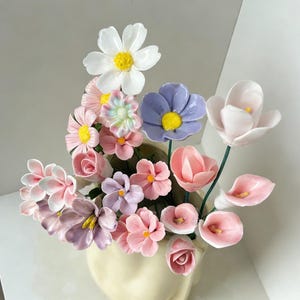 Puede incluir: Un jarrón de cerámica con forma de rostro contiene un colorido arreglo de flores artificiales. Las flores incluyen margaritas blancas, rosas rosas y flores moradas y rosas. El jarrón es de color crema claro.