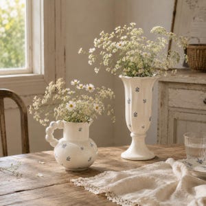 May include: Two white ceramic vases with blue floral patterns, filled with white flowers, sit on a wooden table. One vase is a pitcher with a looped handle, and the other is a tall, pedestal vase. A white lace runner and a glass of water are also on the table.