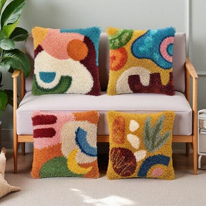 Pode incluir: Quatro almofadas decorativas com designs abstratos. Cada almofada apresenta uma paleta de cores diferente, incluindo rosa, azul, verde e amarelo. As almofadas são colocadas em um sofá claro com braços de madeira.