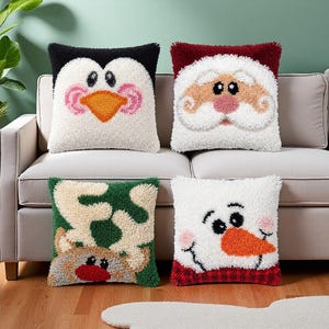 Op de afbeelding: Vier vierkante kussens met getextureerde punch needle ontwerpen. Eén toont een pinguïn, één een kerstmangezicht, één een rendier met gewei op een groene achtergrond en één een sneeuwpop met een oranje neus en een rode ruitjesrand.