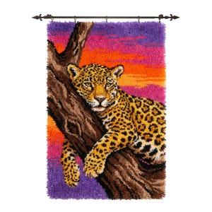 Puede incluir: Un tapiz rectangular de ganchillo que muestra un leopardo descansando sobre un tronco de árbol marrón. El fondo presenta un degradado vibrante de atardecer en tonos morado, naranja y rosa. La pieza cuelga de una barra de metal oscuro con remates.