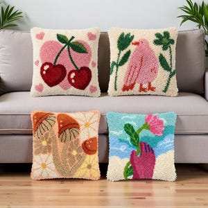 Puede incluir: Cuatro cojines cuadrados con diseños texturizados de aguja mágica. Uno presenta cerezas rojas sobre un corazón rosa, un pájaro rosa con plantas verdes, setas marrones con flores amarillas y una mano sosteniendo una flor rosa sobre azul.