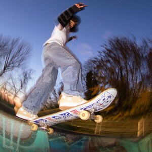 May include: A skateboarder in mid-air, performing a trick at a skate park. The skateboard has a white deck with blue and red graphics. The skater wears a white t-shirt, blue jeans, and tan sneakers. The background is a blurred view of trees and a blue sky.